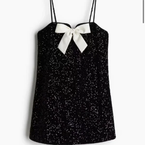 Svart glittrig klänning med rosett - Snygg svart glittrig klänning med en vit rosett framtill. Klänningen har tunna axelband och en elegant design som passar perfekt för festliga tillfällen. Prislappen är kvar!