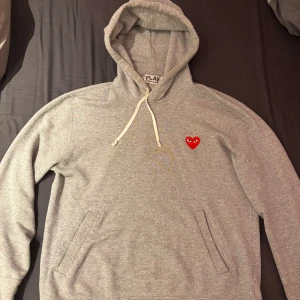 Grå hoodie från Comme des Garçons - Säljer en stilren grå hoodie från Comme des Garçons med det ikoniska röda hjärtat på bröstet. Tröjan har en bekväm passform . Perfekt för en avslappnad look. inga defekter alls köpt på cettire