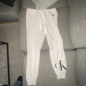 Gråa mjukisbyxor från Calvin Klein - Säljer ett par vita mjukisbyxor från Calvin Klein med snörning i midjan och en stor CK-logga på benet. Perfekta för en avslappnad stil.