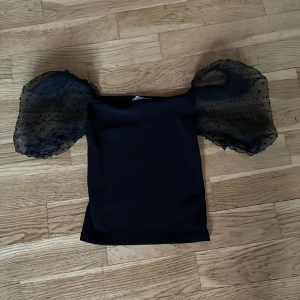 Svart topp med puffärmar från Amisu - Snygg svart topp från Amisu med puffiga, transparenta ärmar dekorerade med små prickar. Perfekt för en trendig look. Toppen har en tight passform och är tillverkad i ett mjukt material.