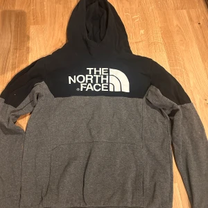 Grå och svart hoodie från The North Face - Säljer en snygg grå och svart hoodie från The North Face med stor logga på bröstet. Tröjan har en bekväm passform och en praktisk känguruficka framtill. Perfekt för kyliga dagar!