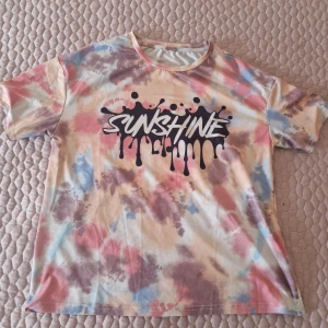 Färgglad t-shirt med tryck - Säljer en färgglad t-shirt med texten 'Sunshine' i svart tryck. T-shirten har ett tie-dye mönster i rosa, blått och beige. Den är kortärmad och har en rund halsringning.