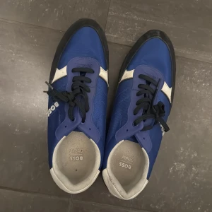 Blå sneakers från Boss - Snygga blå sneakers från Boss med vita detaljer och svart sula. Skorna har snörning och en stilren design som passar perfekt för en avslappnad look.