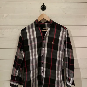 Rutig skjorta från Burberry - Snygg rutig skjorta från Burberry England i svart, vit och röd. Skjortan har långa ärmar och knappar framtill. Perfekt för en fresh look. Skjortan är perfekt till en fin sommar dag när det är fint väder ute t.ex på midsommar men även en helt vanlig dag. Skjortan har ett broderat Burberry märke på bröstet. Skjortan är ej äkta och det är därför jag säljer den för ett rimligt pris, skjorta är i väldigt bra skick använd fåtal gånger. Ni får gärna höga av er om ni undrar något! Pris ej hugget i sten!