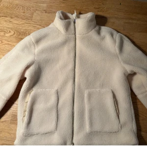 Beige teddyjacka - Mysig beige teddyjacka med dragkedja och hög krage. Jackan har två stora fickor framtill och är perfekt för kyliga dagar. Den är tillverkad i ett mjukt och fluffigt material som ger en skön känsla. Storlek s men passar även xs.