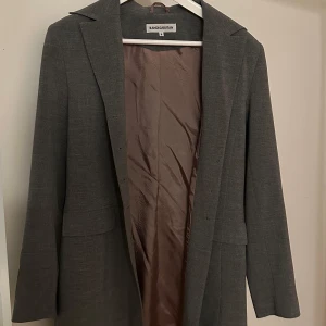 Grå kappa från Randigarutan - Elegant grå kappa från Randigarutan med klassisk design. Kappan har långa ärmar och en stilren krage. Perfekt för höst- och vårsäsongen. Den är tillverkad i ett mjukt material som ger en bekväm passform.