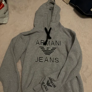 Grå hoodie från Armani Jeans - Snygg grå hoodie från Armani Jeans med svart logotyp på bröstet. Tröjan har en klassisk känguruficka och justerbar huva med svarta snören. Perfekt för en avslappnad stil. Passar både xs och s.