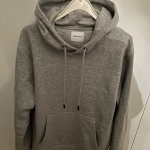 Grå hoodie från Jack & Jones - Säljer en stilren grå hoodie från Jack & Jones. Den har en klassisk design med en stor ficka framtill och justerbar dragsko i huvan. Perfekt för en avslappnad look. Köpt på nypris för 599 i butik