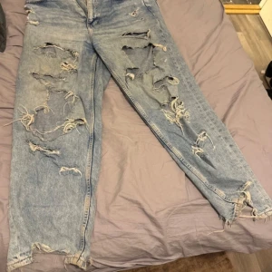 Blå slitna jeans - Säljer ett par blå jeans med en cool, sliten look. De har flera revor och slitningar som ger en edgy stil. Perfekta för dig som gillar en avslappnad och trendig outfit. 