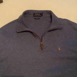 Blå tröja med dragkedja från Ralph Lauren - Snygg blå tröja från Ralph Lauren med halv dragkedja och klassisk krage. Den har ett broderat logotypmärke på bröstet och är perfekt för en stilren look.