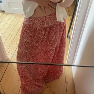 Röd paisleymönstrad långkjol - Säljer en vacker röd långkjol med paisleymönster och fina pärldetaljer vid midjan. Kjolen har en lös passform och är perfekt för en bohemisk stil. Den är tillverkad i ett lätt och luftigt material som passar bra för varmare väder.