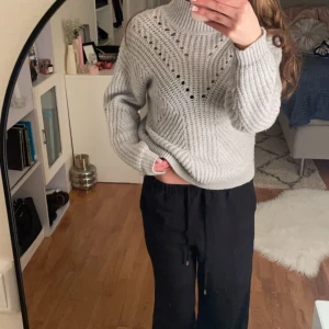 Grå stickad tröja - Säljer en mysig grå stickad tröja med ett dekorativt hålmönster framtill. Tröjan har en hög krage och långa ärmar, perfekt för kyligare dagar. Den är både stilren och bekväm, idealisk för en avslappnad look.