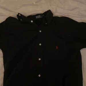 Svart skjorta från Polo - Snygg svart skjorta från Polo i 100% bomull. Den har en klassisk design med knappar och en liten broderad logga på bröstet. Perfekt för en stilren look. Storleken är L men sitter Max som en M