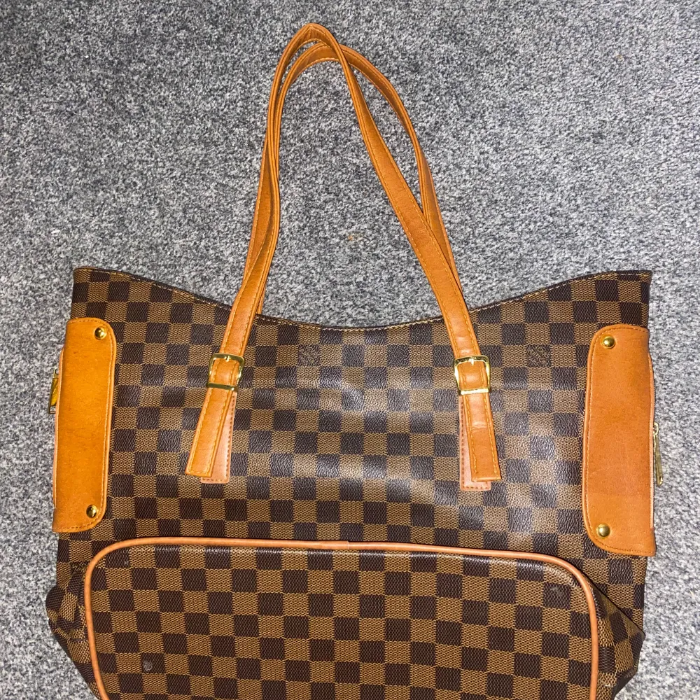 Snygg axelväska med klassiskt rutigt mönster i brunt och beige. Väskan har bruna handtag och detaljer i skinn samt en guldfärgad platta framtill. Perfekt för att bära med sig allt man behöver.. Laukut & Käsilaukut.