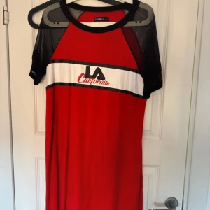 Röd klänning från FB Sister - Trendig röd klänning med svart och vit text 'LA California' framtill. Klänningen har svarta meshdetaljer på ärmarna och en rund halsringning. Perfekt för en avslappnad stil.