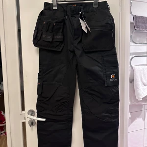 Svarta arbetsbyxor från Worksafe - Säljer ett par svarta arbetsbyxor från Worksafe med flera praktiska fickor och förstärkta knän. Byxorna är designade för komfort och funktionalitet, perfekta för aktiva personer. De har en stretchig passform och är tillverkade för att hålla hela dagen.