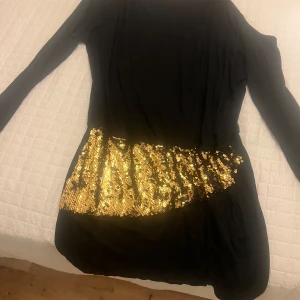 Svart klänning med guldpaljetter - Snygg svart klänning med glittrande guldpaljetter över midjan. Klänningen har långa ärmar och en tight passform som framhäver figuren. Perfekt för en festlig kväll!