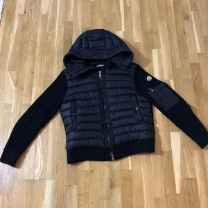 Svart Moncler Cardigan - Snygg svart quiltad jacka från Moncler med huva och dragkedja. Jackan har långa ärmar och ribbade detaljer vid ärmslut och nederkant. Perfekt för kyligare dagar.