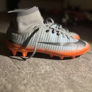 Nike Mercurial fotbollsskor - Snygga Nike Mercurial fotbollsskor i grått och orange med snörning och hög ankelkrage för extra stöd. Perfekta för snabba rörelser på planen.