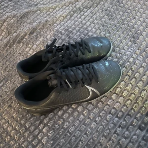 Svarta fotbollsskor från Nike - Snygga svarta fotbollsskor från Nike med en diskret silverdetalj på sidan. Skorna har snörning och en slitstark sula, perfekt för spel på olika underlag.