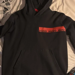 Svart hoodie från Hugo - Säljer en svart hoodie från Hugo med röd logga på bröstet. Tröjan har en klassisk känguruficka och en bekväm huva. Perfekt för en avslappnad stil.