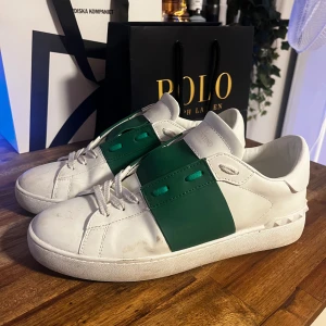 Valentino sneakers med grönt band - Snygga vita Valentino sneakers med ett brett grönt band över snörningen. Skorna har en stilren design med en platt sula och diskreta detaljer. Perfekta för en trendig look. Vet inte min det är o***nal