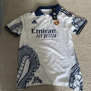 Vit Real Madrid fotbollströja med drakmönster - Snygg vit Real Madrid fotbollströja från Adidas med ett unikt blått drakmönster. Tröjan har korta ärmar och en klassisk krage. Perfekt för fans som vill sticka ut på läktaren eller planen. Officiellt licensierad produkt med Aeroready-teknologi för optimal komfort.