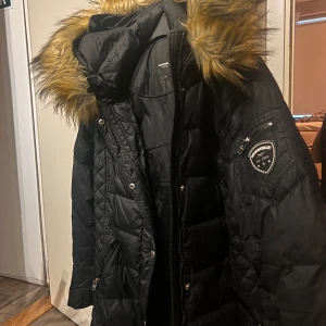 Svart dunjacka med päls från Rockandblue - Snygg svart dunjacka från Rockandblue med quiltad design och avtagbar huva med fuskpäls. Jackan har dragkedja och knappar framtill samt en stilren patch på ärmen. Perfekt för kyliga dagar!