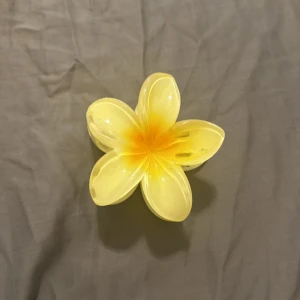 Gul blomformad hårklämma - Söt hårklämma i form av en blomma med fem kronblad. Klämman är i en ljusgul färg med en orange nyans i mitten, perfekt för att ge en somrig touch till din frisyr.