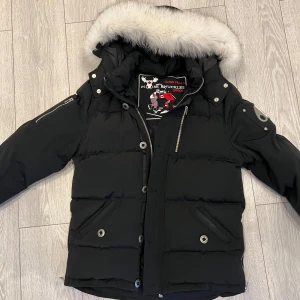 Svart Moose Knuckles jacka med vit päls  - Snygg svart dunjacka från Moose Knuckles med pälskantad huva med vit päls. Jackan har dragkedja och knappar framtill samt flera fickor. Perfekt för kalla vinterdagar.