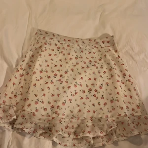 Blommig kjol med volang - Charmig kortkjol i vitt med ett sött blommönster i rosa och grönt. Kjolen har en volangdetalj längst ner som ger en lekfull touch. Perfekt för varmare dagar.