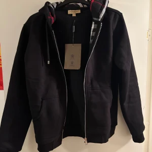 Svart hoodie från Burberry - Säljer en stilren svart hoodie från Burberry med dragkedja och justerbar huva. Den har ett klassiskt rutigt mönster på insidan av huvan och är tillverkad i mjuk bomull. Perfekt för en avslappnad och trendig look. Aldrig använd och är storlek M