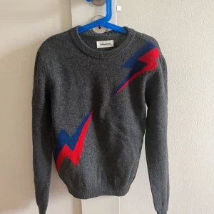 Grå Zadig & Voltaire tröja med blixtar - Snygg grå tröja från Zadig & Voltaire med blixtmotiv i rött och blått. Perfekt skick. Tröjan är långärmad och tillverkad i ull, perfekt för kyligare dagar. Storlek 12/14 men passar mig som brukar ha S/XS. Pris kan diskuteras💕💕💕