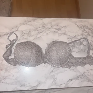 Vit spets-bh - Elegant grå bh med spetsdetaljer och en liten dekorativ rosett framtill. Den har justerbara axelband och en klassisk hakknäppning i ryggen. Storlek: 80A