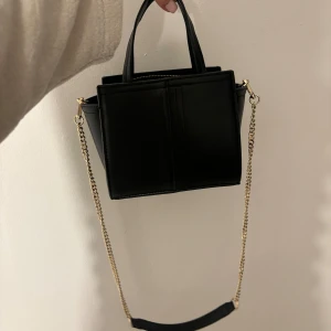Zara Svart handväska med kedja - Snygg svart handväska med guldfärgad kedja och avtagbar axelrem. Perfekt för att ge en elegant touch till din outfit. Väskan har en stilren design och är lätt att matcha med olika stilar. Rymmer mkt, knappt använd