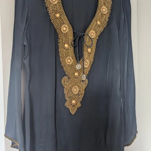 Svart tunika/kaftan med guldbroderi - Snygg tunika eller kaftan med vackra pärlbroderier i guld. Ledig modell i svart, med v-ringning och snörning framtill. Långa ärmar som avslutas med pärlbroderi i guld.