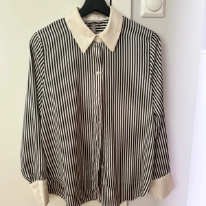 Randig skjorta från H&M - Säljer en superfin randig skjorta från H&M. Den har en klassisk svart och vit randig design med en kontrasterande vit krage och manschetter. Skjortan är långärmad och har knappar framtill. Perfekt för både vardag och fest!