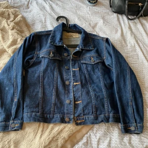 Blå jeansjacka från Wrangler - Säljer en klassisk blå jeansjacka från Jeanswear. Den har en snygg och tidlös design med knappar framtill och två bröstfickor med knappstängning. Jackan är i bra skick och perfekt för både vår och höst. Passar till nästan allt i garderoben!