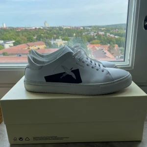 Vita sneakers från Axel Arigato - Säljer ett par stilrena vita sneakers från Axel Arigato. Skorna har en snygg svart detalj på sidan med en fågel. De är i skinn och har klassisk snörning. Perfekta för en casual look! Model ”Clean 90”