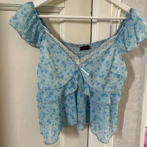 Blå blommig offshoulder blus - Söt blå blommig offshoulder blus med volanger och spetsdetaljer. Perfekt för sommardagar och har en liten rosett framtill. Den är luftig och lätt, vilket gör den idealisk för varma väder.
