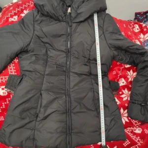 Svart dunjacka med huva - Snygg svart dunjacka med dragkedja och stor huva. Jackan har långa ärmar och justerbara remmar i midjan för en bättre passform. Perfekt för kyliga dagar.