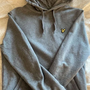 Grå hoodie från Lyle & Scott - Snygg grå hoodie från Lyle & Scott med det klassiska logotypmärket på bröstet. Perfekt för en avslappnad stil med justerbar huva och långa ärmar. Passar bra till både jeans och joggers.