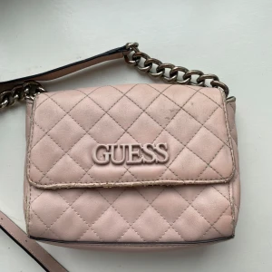 Rosa äkta guess väska - Snygg rosa axelväska från Guess med quiltat mönster och kedjedetaljer. Väskan har en justerbar axelrem och ett stilrent Guess-emblem på framsidan.  Väskan har använts några gånger så den är lite sliten runt kanterna och på kedjan. Pruta gärna❤️
