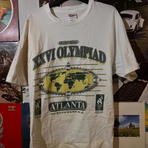 Vit single stitch vintage t-shirt från Hanes - Klassisk vit t-shirt från Hanes med tryck från XXVI Olympiad i Atlanta 1996. Designen inkluderar en världskarta och text i grönt och gult. Perfekt för den som älskar retro sportmode!