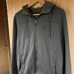 Grå huvtröja från Hugo Boss - Säljer en stilren grå huvtröja från Hugo Boss med dragkedja och broderad logga på bröstet. Tröjan har långa ärmar och en justerbar huva med dragsko. Perfekt för en avslappnad look. Limiterad färg som är slutsåld överallt, köpt i milano 2018.