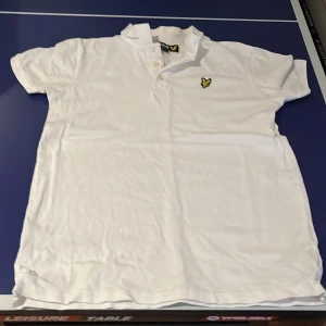 Vit pikétröja från Lyle & Scott - Säljer en klassisk vit pikétröja från Lyle & Scott med korta ärmar och broderad logga på bröstet. Perfekt för en stilren look.
