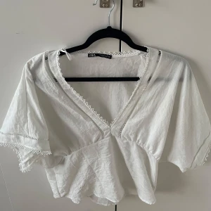 Vit blus från Zara - Säljer en söt vit blus från Zara med v-ringning och spetsdetaljer längs kanterna. Blusen har korta ärmar och en luftig passform.