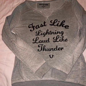 Grå tröja från True Religion - Säljer en grå tröja från True Religion med texten 'Fast Like Lightning, Loud Like Thunder' i svart glitter. Tröjan har långa ärmar och en bekväm passform. Perfekt för en avslappnad stil.