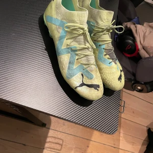Fotbollsskor från Puma - Snygga fotbollsskor från Puma i en fräsch gul och turkos färgkombination. Skorna har snörning och en strumpliknande passform för extra komfort. Perfekta för fotbollsplanen med sina greppvänliga dobbar.