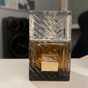 Khamsah Khamrah Parfym från Lattafa - Elegant parfymflaska med ett unikt mönster i glaset. Flaskan har en gyllene etikett och en djup bärnstensfärgad vätska. Förpackningen är lyxig med bruna och guldiga detaljer. Perfekt för den som söker en sofistikerad doftupplevelse.